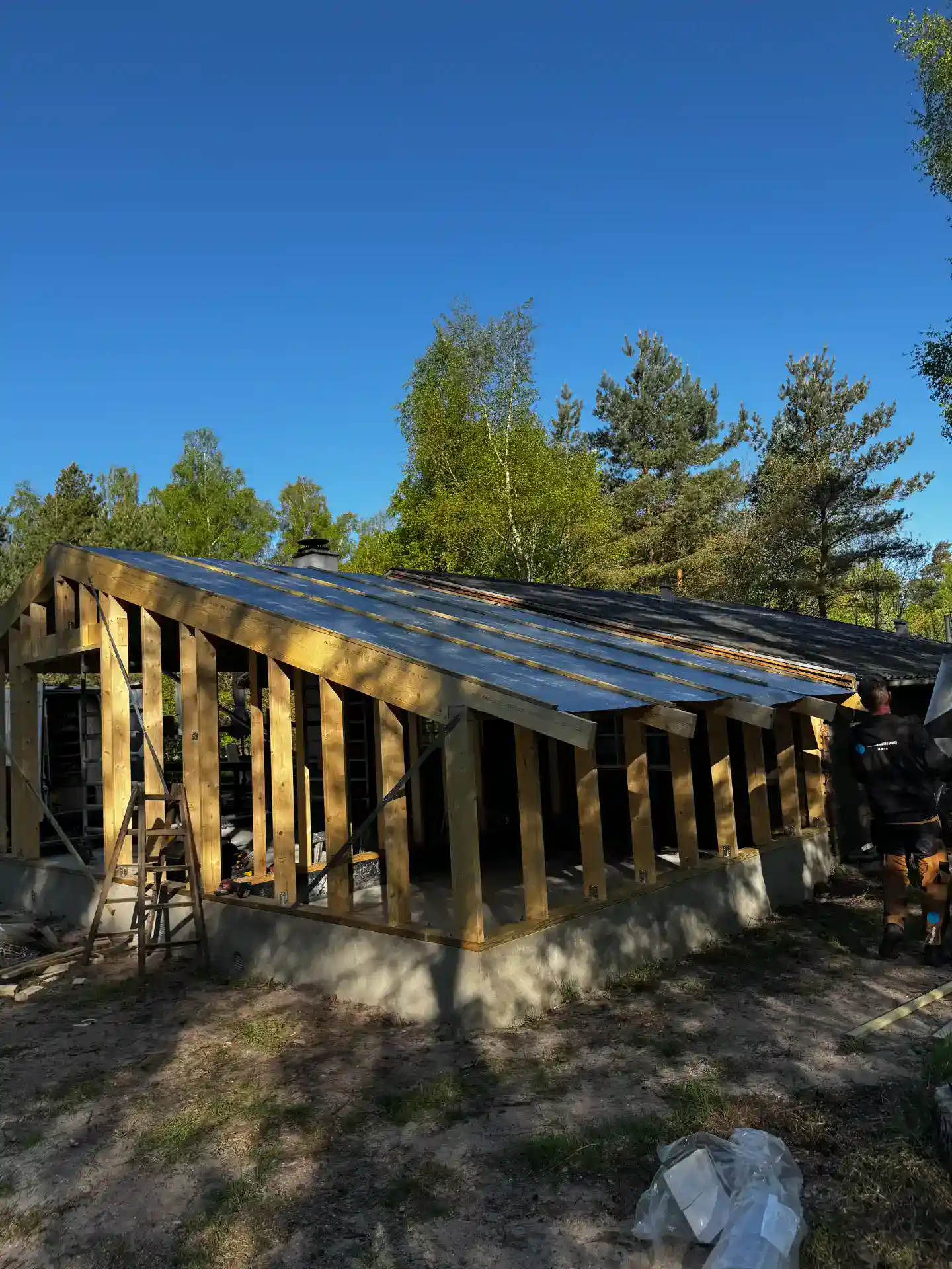 Træværk under opførsel til hus med ståltag og omgivelser af skov.